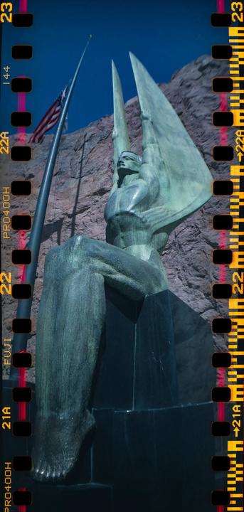 A large bronze statue of a man with wings sits on a pedestal next to a flagpole with an American flag against a rocky background.