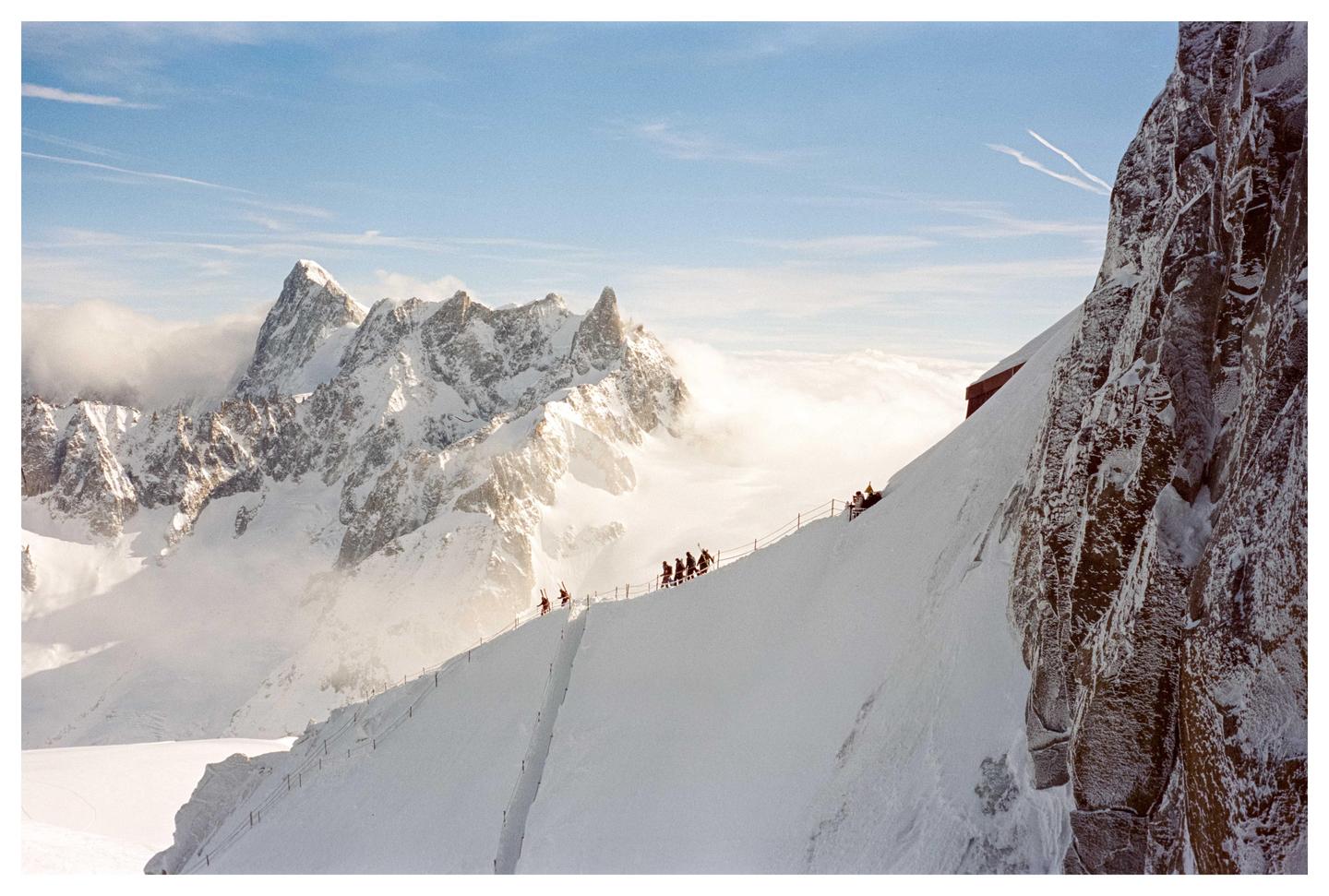 A group of people walk along a snowy mountain ridge with a steep drop-off and jagged peaks in the background.