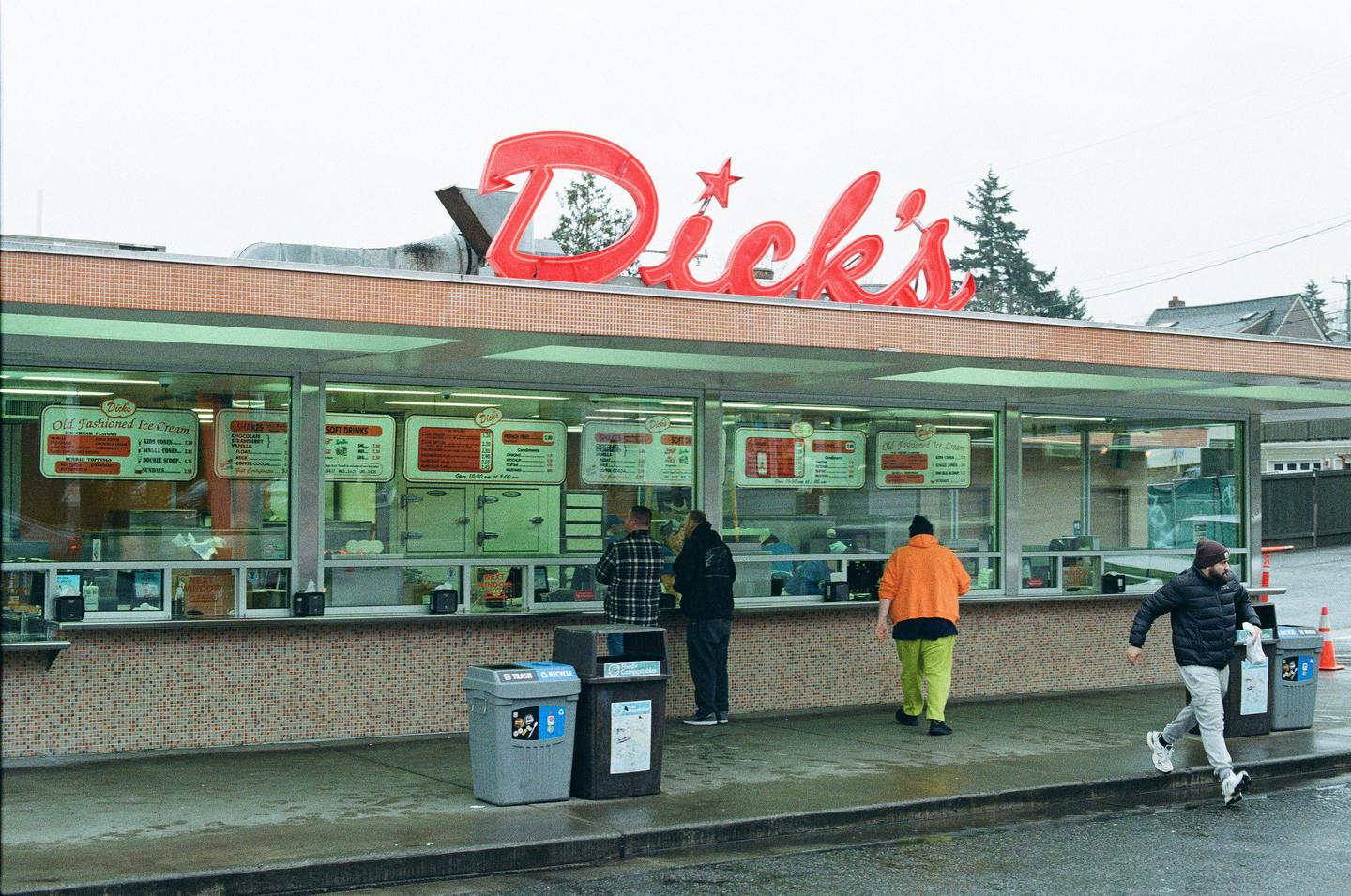 A group of men are standing outside a fast food restaurant with a red sign that says "Dick's" on a cloudy day.
