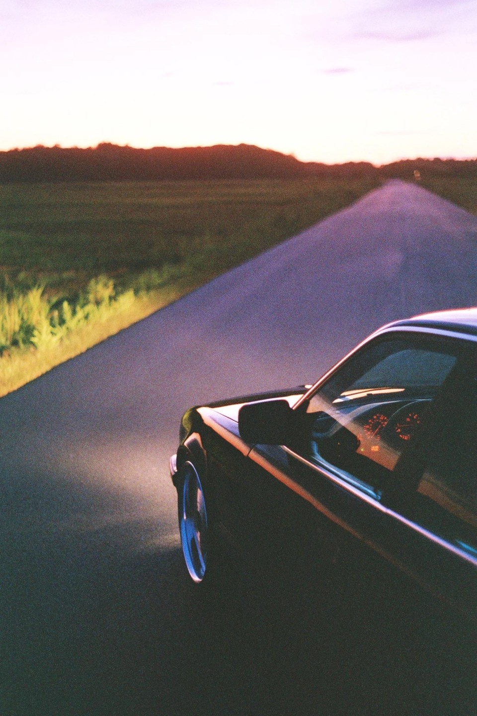 A car drives down a road at dusk, with its headlight illuminating the grassy shoulder.