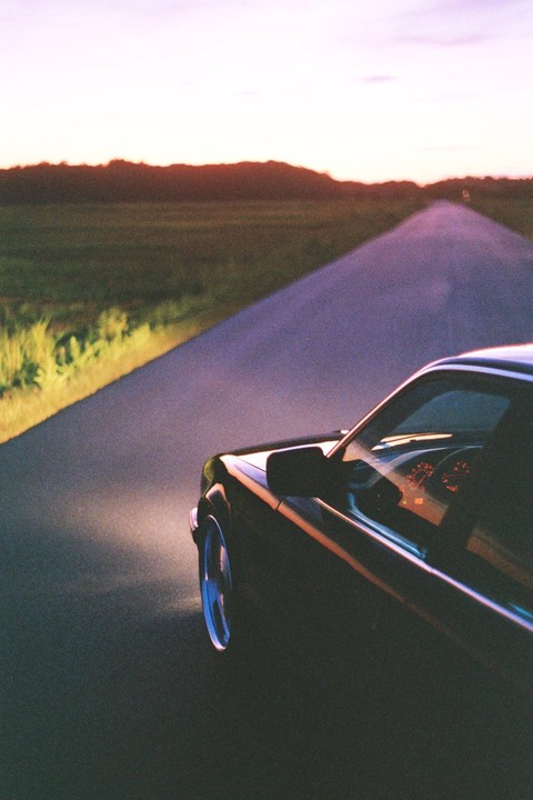 A car drives down a road at dusk, with its headlight illuminating the grassy shoulder.