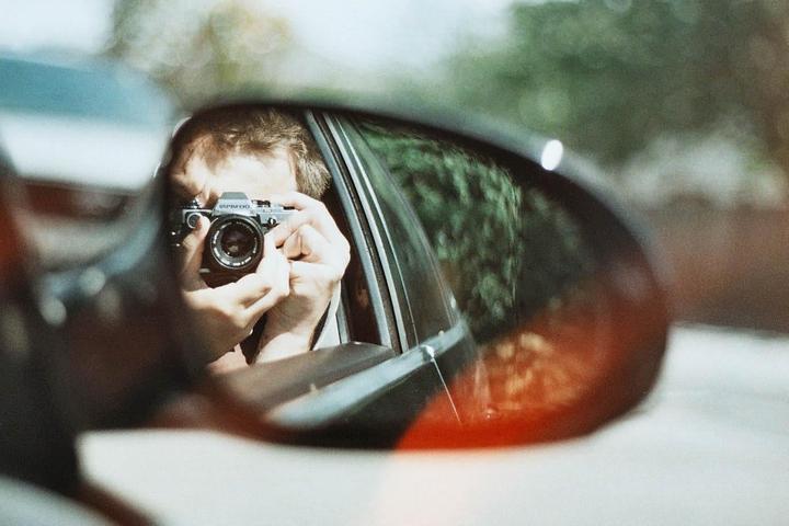 A man is visible in a car's side mirror, holding a camera to take a self-portrait.