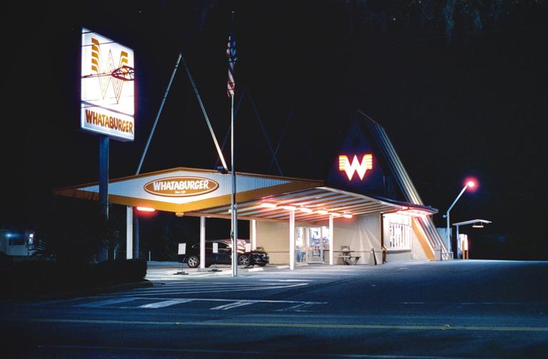 A Whataburger restaurant is illuminated at night with a car parked under its drive-thru canopy.
