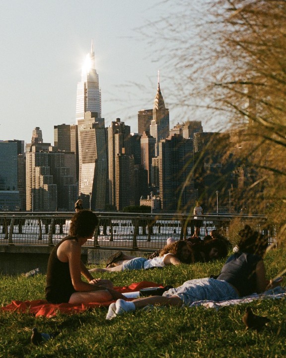 People relax on a grassy bank overlooking a city skyline with a prominent skyscraper reflecting sunlight.