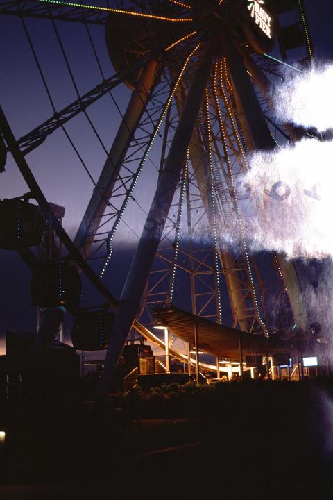 A ferris wheel with illuminated spokes stands against a dark blue sky, with a bright light flare on the right side of the image.