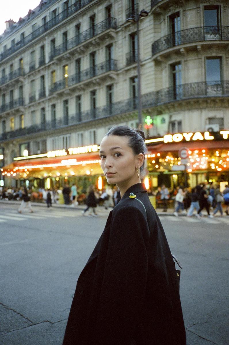 A woman in a black coat looks over her shoulder on a city street with a cafe and building in the background.