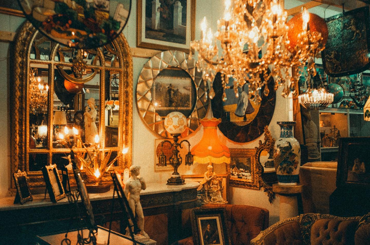 A cluttered room is filled with antique furniture, mirrors, lamps, and chandeliers.