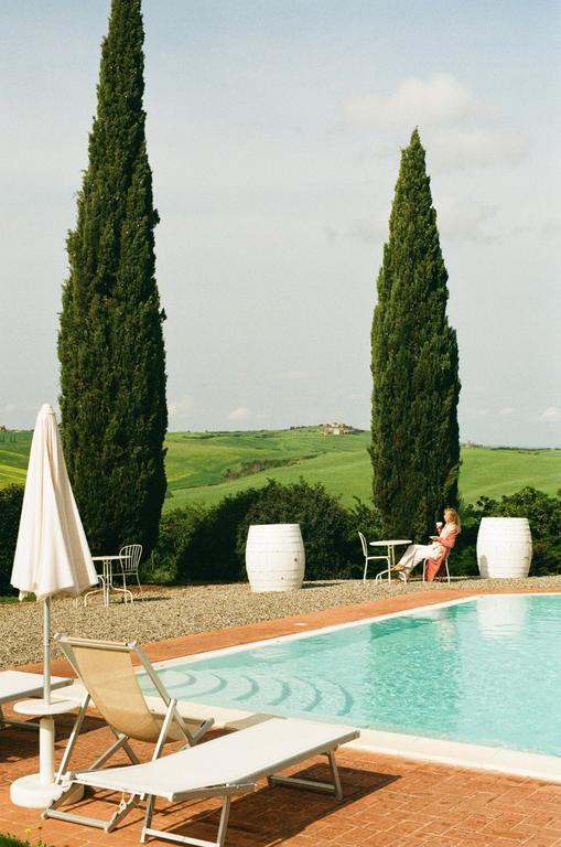 A woman sits at a table next to a swimming pool with cypress trees and rolling hills in the background.