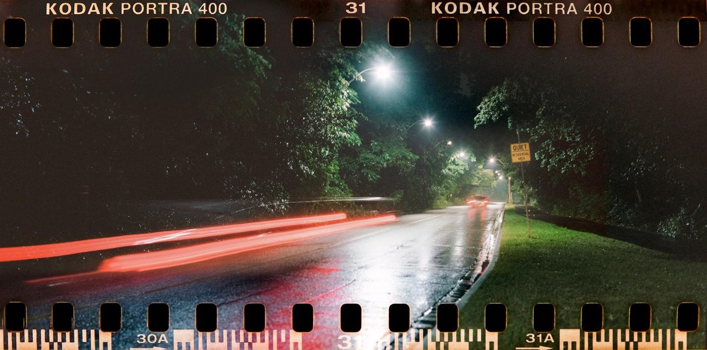 A wet road at night is illuminated by streetlights, with red light trails from a vehicle passing by.
