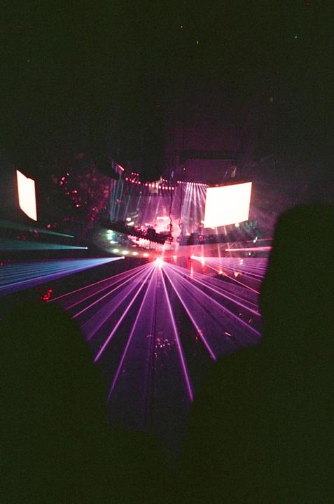 Purple laser beams illuminate a concert stage with a large screen and performers.