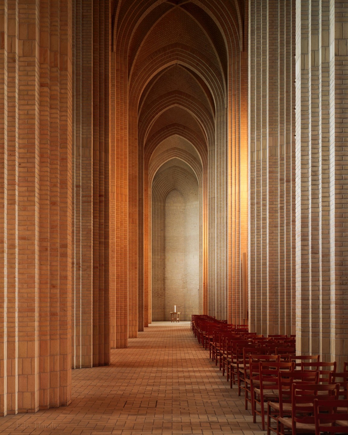 A long hallway with brick columns and arches leads to a single lit candle on a small table, with rows of red chairs on the right.