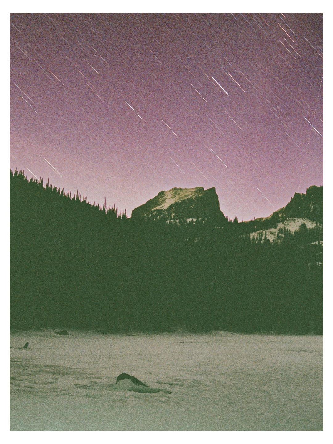 A long exposure captures star trails over a snow-covered landscape with mountains and a forest.