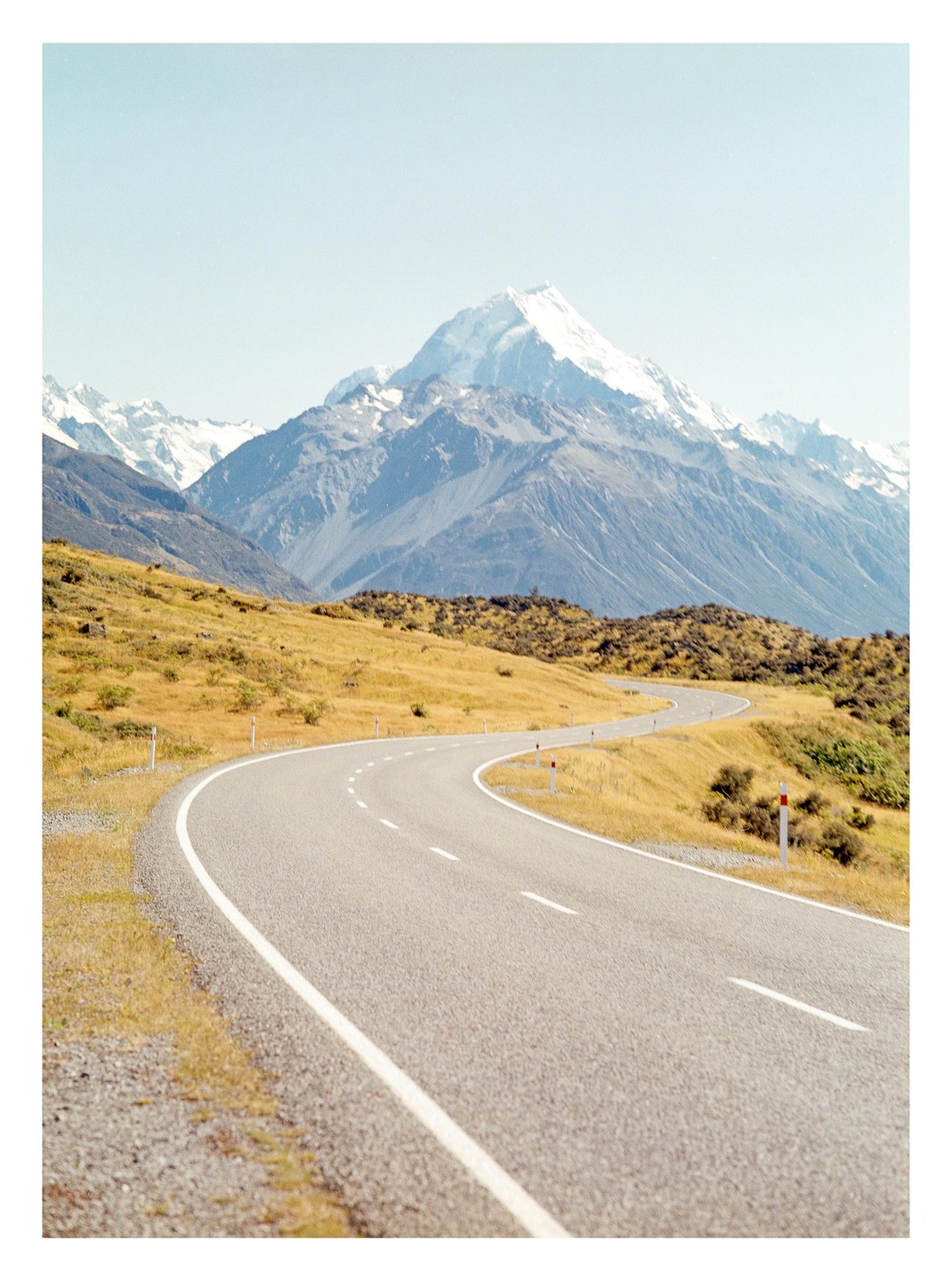 A winding road leads through a golden field towards a snow-capped mountain under a clear sky.