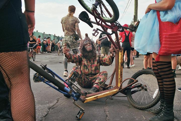 A person in a gorilla mask and camouflage jumpsuit sits on the ground next to a bicycle, surrounded by other people.
