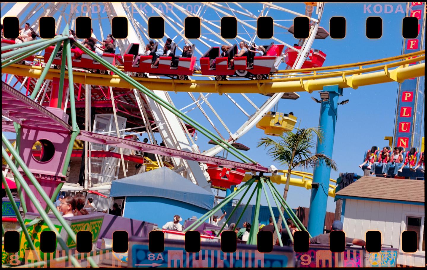 A roller coaster with people riding in red cars is visible against a clear blue sky at an amusement park.