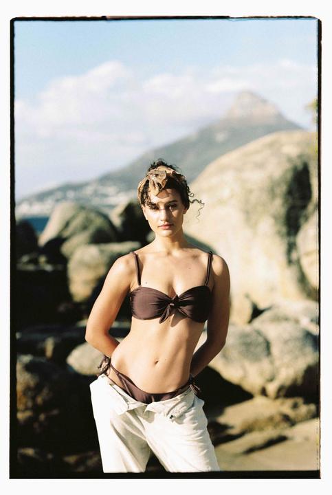 A woman in a bikini top and unbuttoned pants stands on a rocky beach with a mountain in the background.