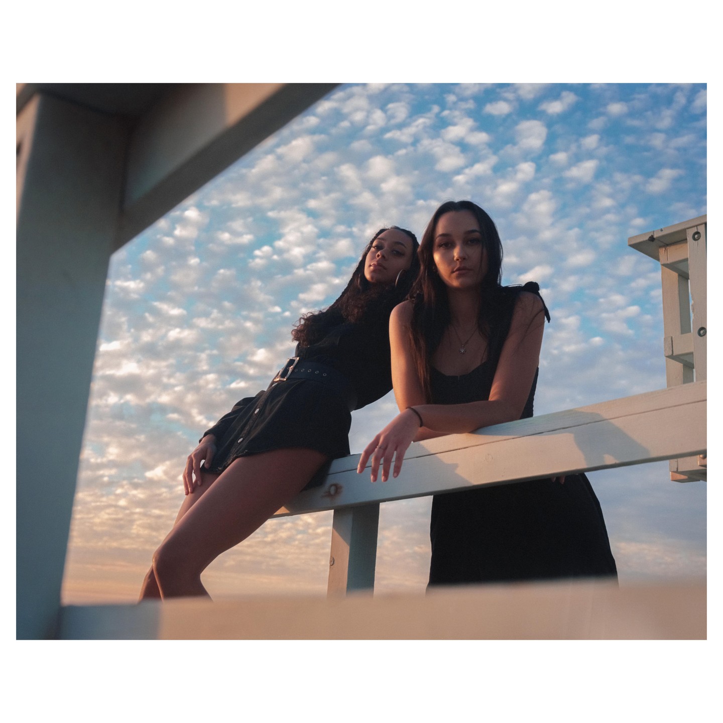 Two women pose on a lifeguard stand against a cloudy sky at sunset.