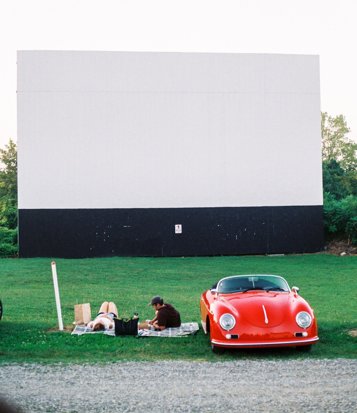 A couple sits on a blanket in front of a large drive-in movie screen with a red vintage car parked nearby.