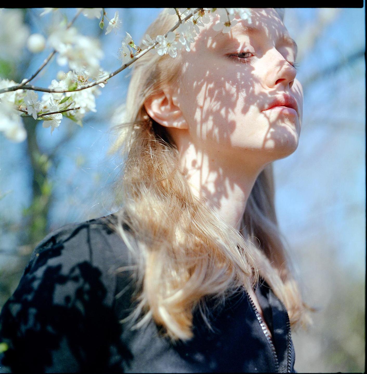 A woman with long blonde hair looks up at the sky, with shadows of leaves on her face and a branch of white blossoms in the foreground.