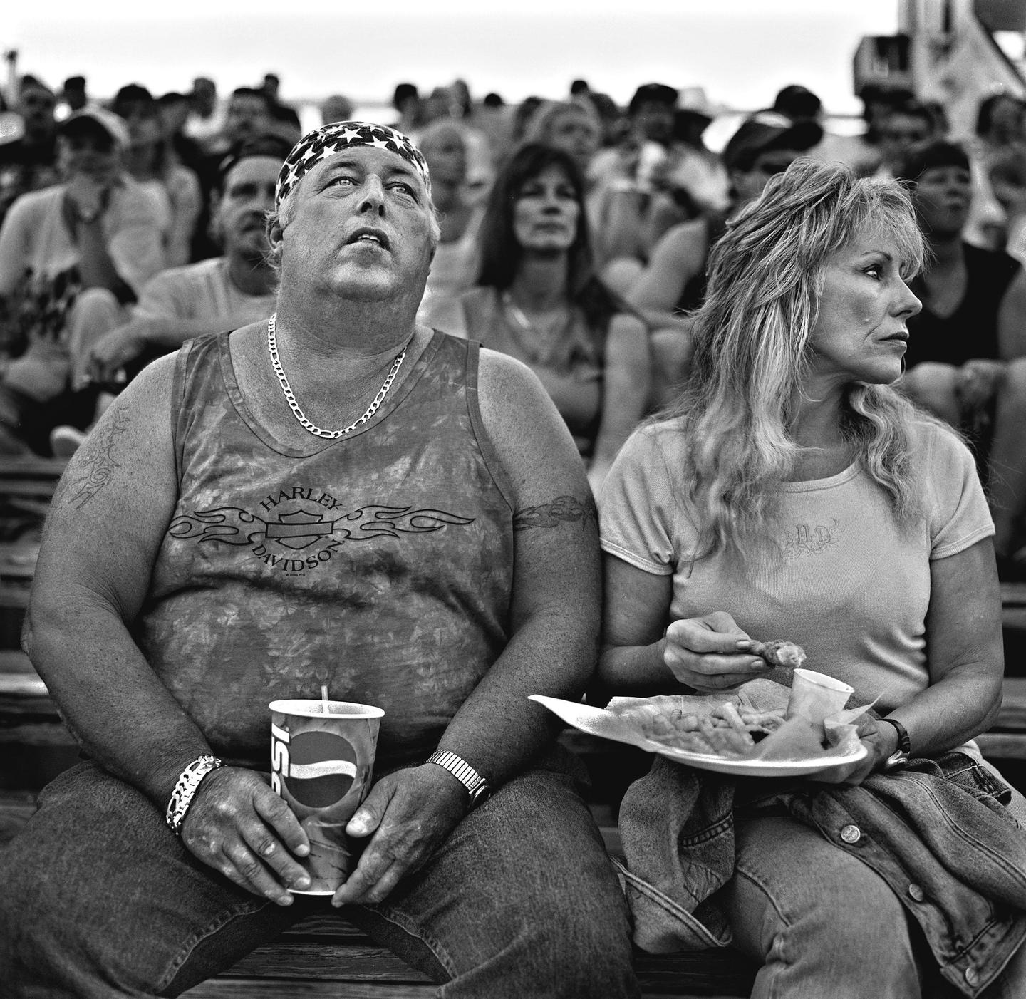 A man and a woman sit in a crowd, the man holding a drink and the woman eating from a paper plate.