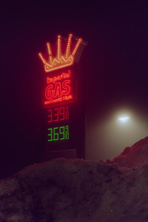 A neon sign for Imperial Gas and Country Store with gas prices is visible at night, with a pile of snow in the foreground.