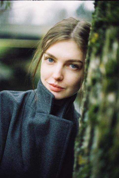 A woman with blue eyes and a dark coat stands next to a tree.