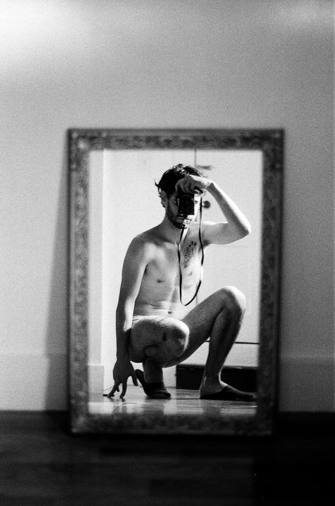 A man crouches in front of a mirror, holding a camera to his face.