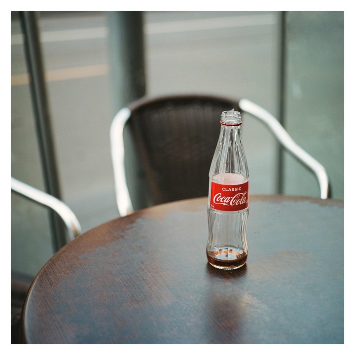 An empty Coca-Cola bottle sits on a round wooden table with a chair in the background.