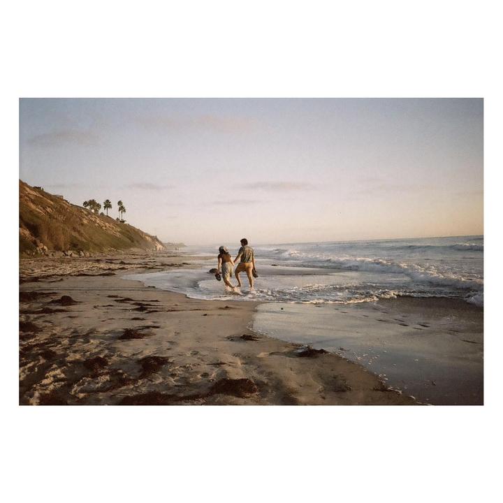 A couple walks along the water's edge on a beach with a cliff and palm trees in the background.