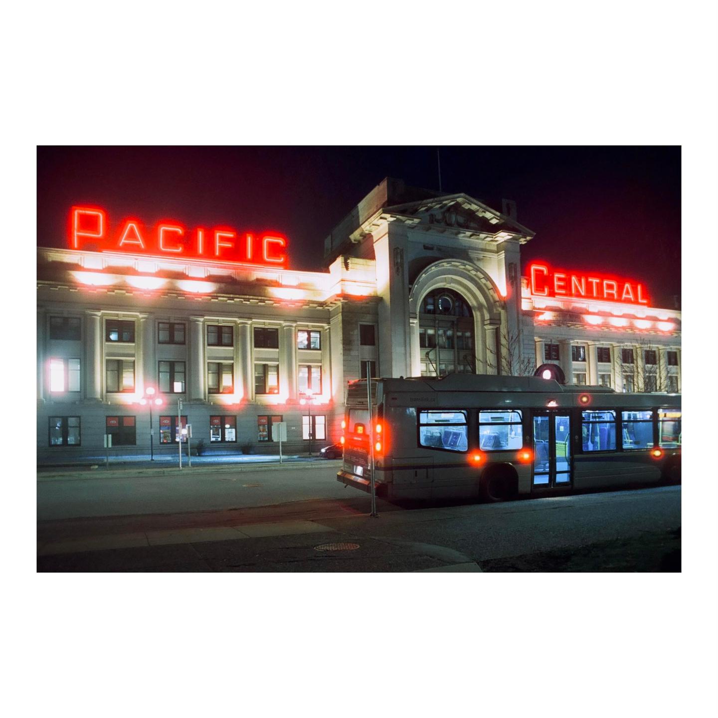 A bus is parked in front of a building with "PACIFIC CENTRAL" written in neon letters on its facade at night.