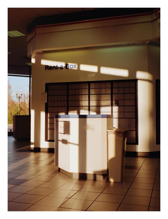 Sunlight streams into a post office lobby with rows of mailboxes and a counter.