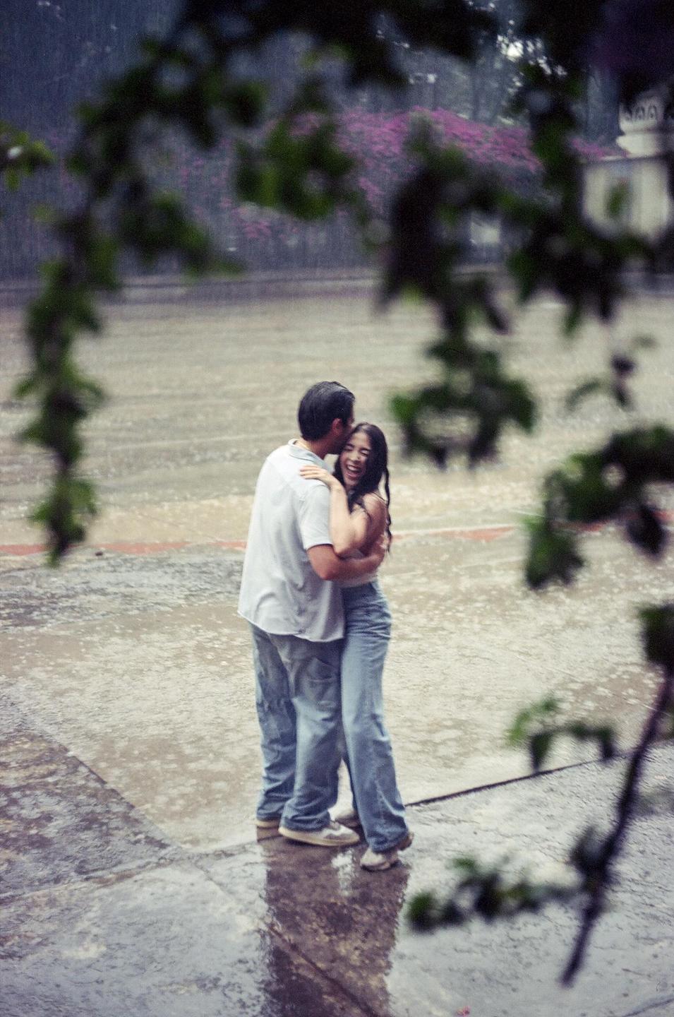 A couple embraces and laughs while standing in the rain on a paved surface, framed by out-of-focus foliage.