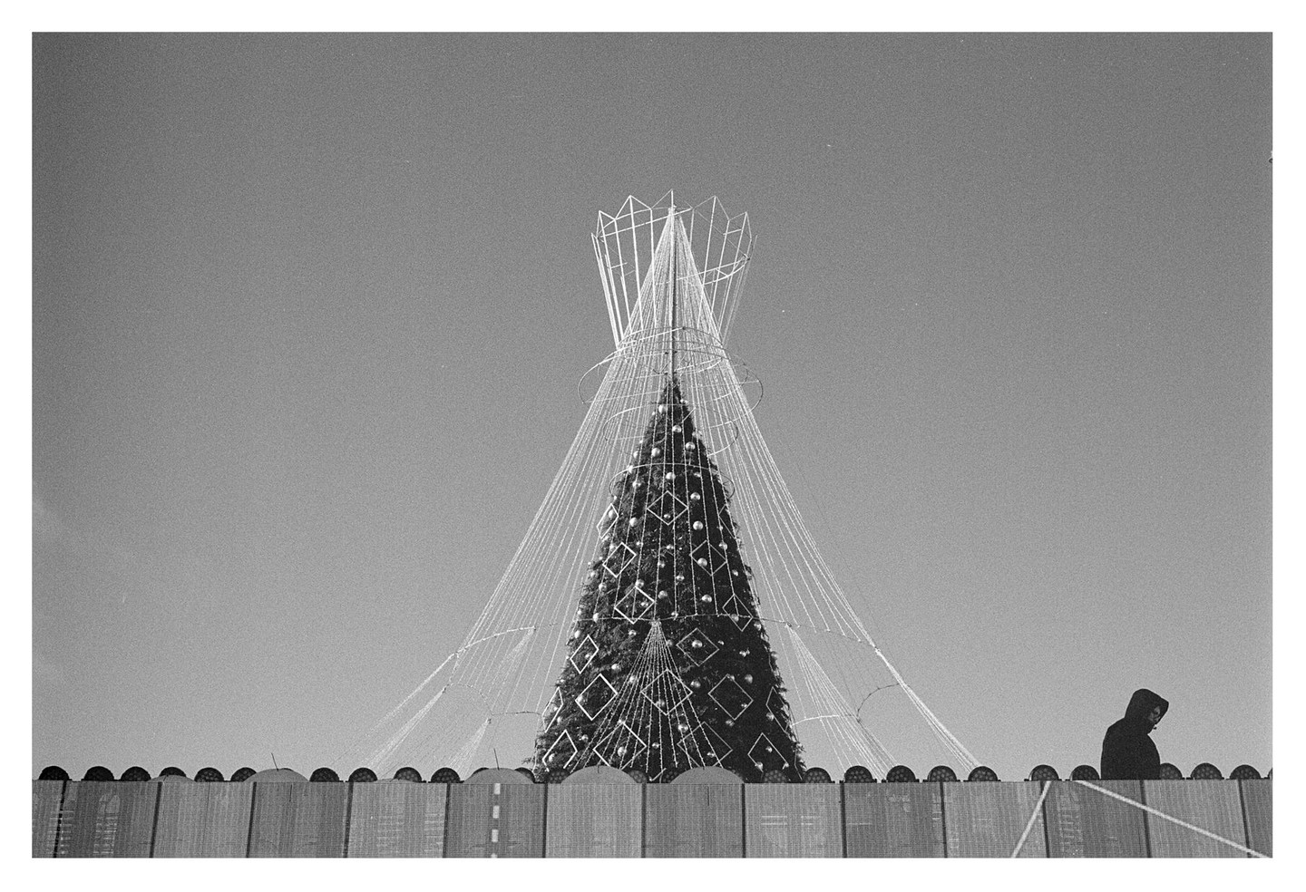 A large Christmas tree decorated with lights and ornaments stands behind a fence, with a person in a hooded jacket visible on the right.