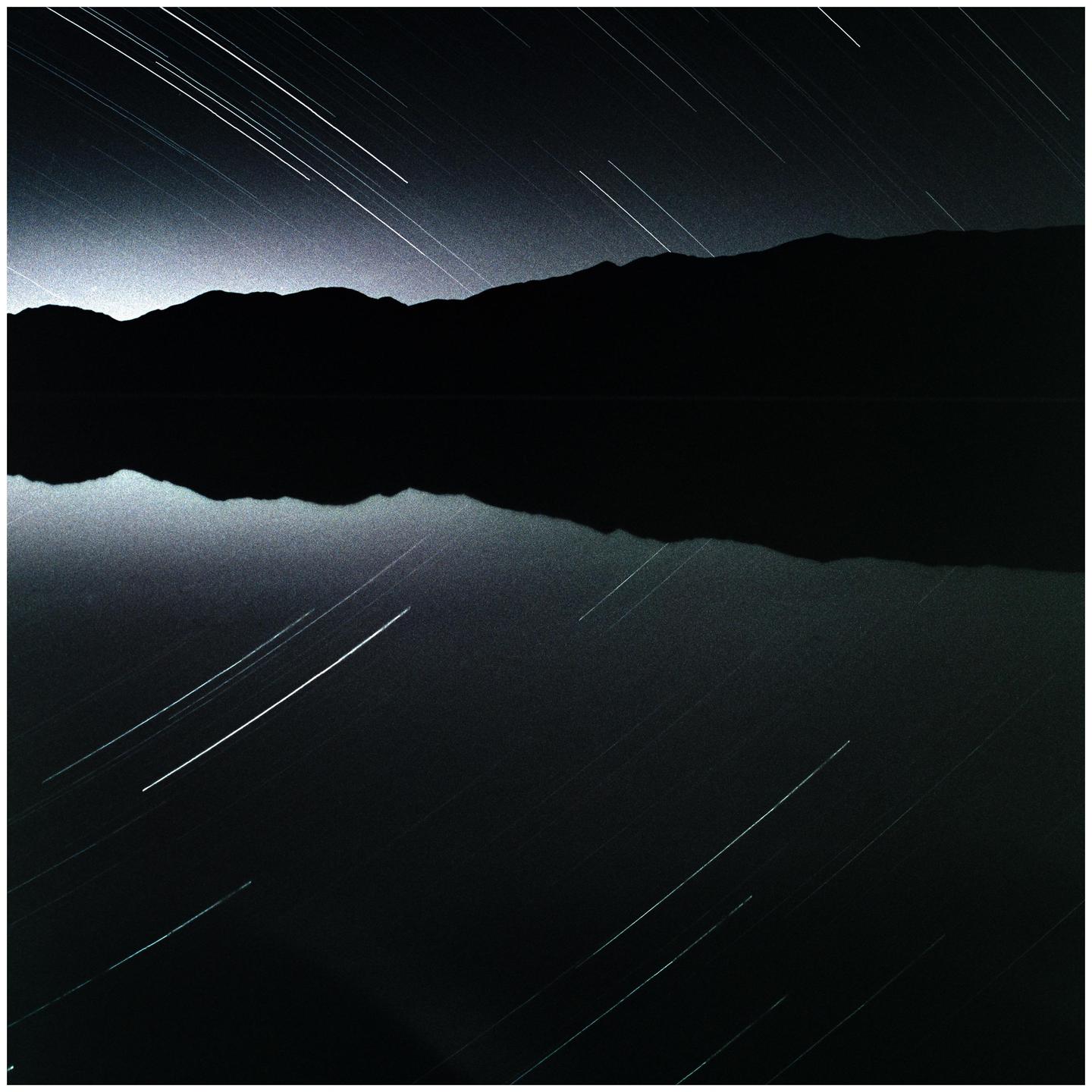 Star trails are visible above a mountain range reflected in a body of water at night.