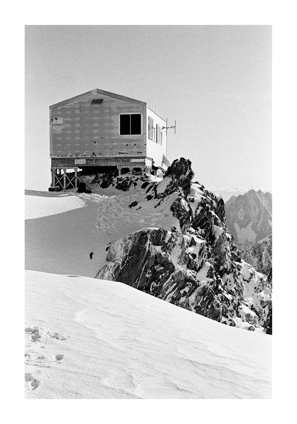 A small building sits atop a snowy, rocky mountain peak with other mountains visible in the distance.
