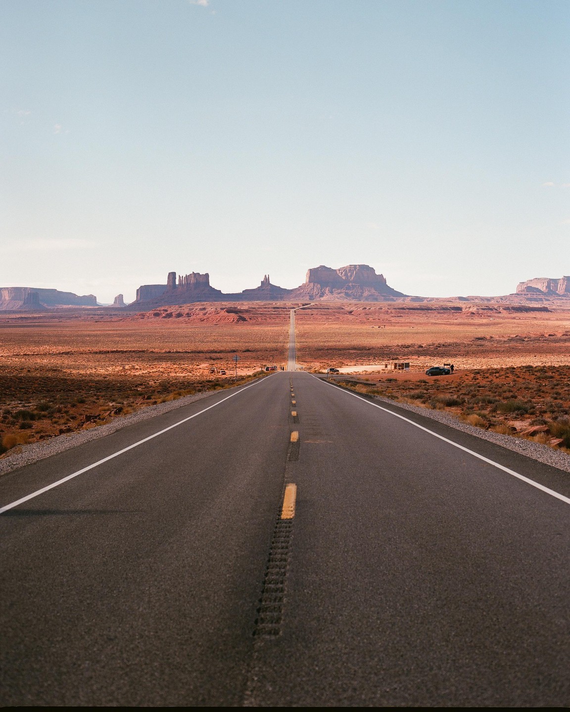 A long, straight road leads through a desert landscape towards distant mesas under a clear sky.