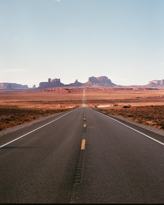 A long, straight road leads through a desert landscape towards distant mesas under a clear sky.
