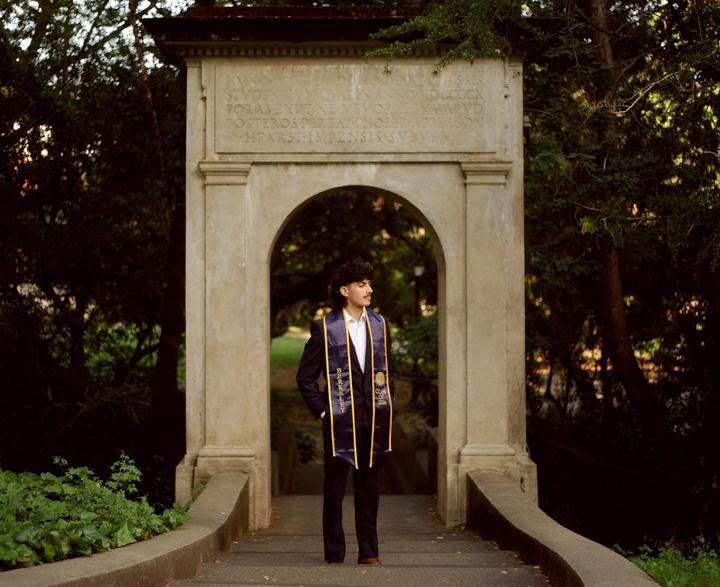 A man in a suit and graduation stole stands on a stone bridge with an archway behind him.