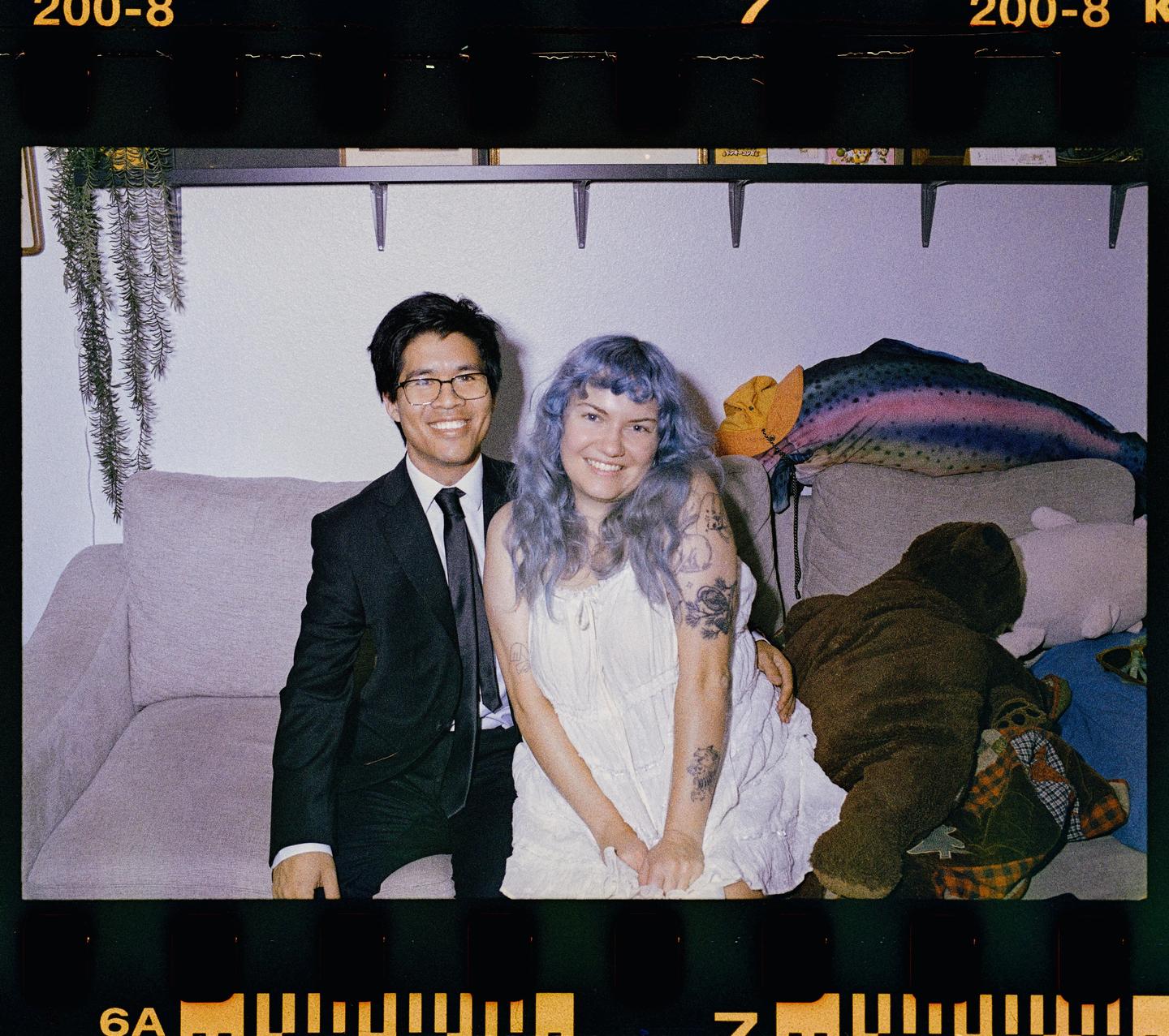 A smiling couple sits on a sofa with a large fish pillow and a teddy bear.