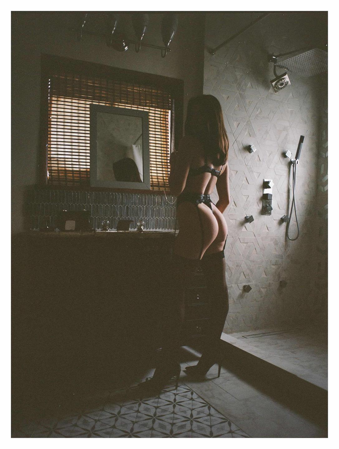 A woman in lingerie stands with her back to the camera in a bathroom with a window and a shower.