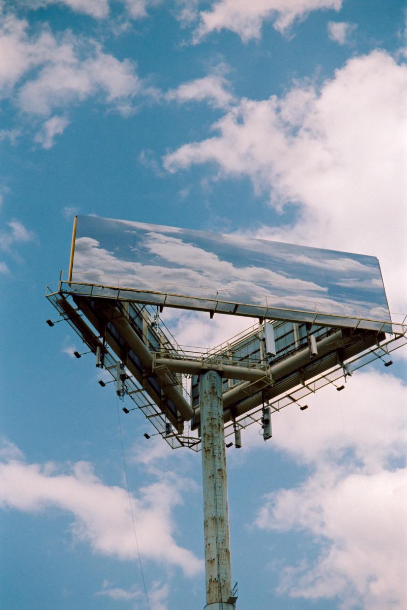 A large billboard with a cloudy sky reflected on its surface stands against a blue sky with white clouds.