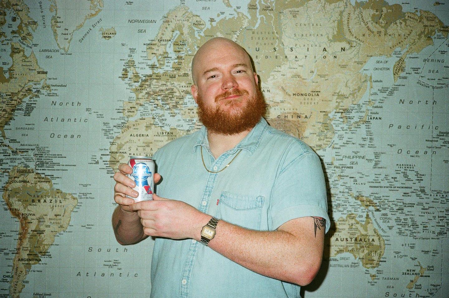 A man with a red beard and a light blue shirt holds a can of beer in front of a world map.