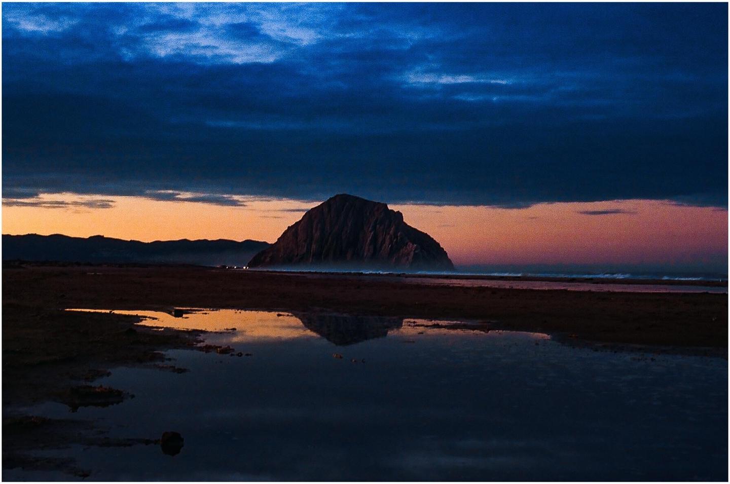A large rock formation is reflected in a pool of water on a beach at dusk.