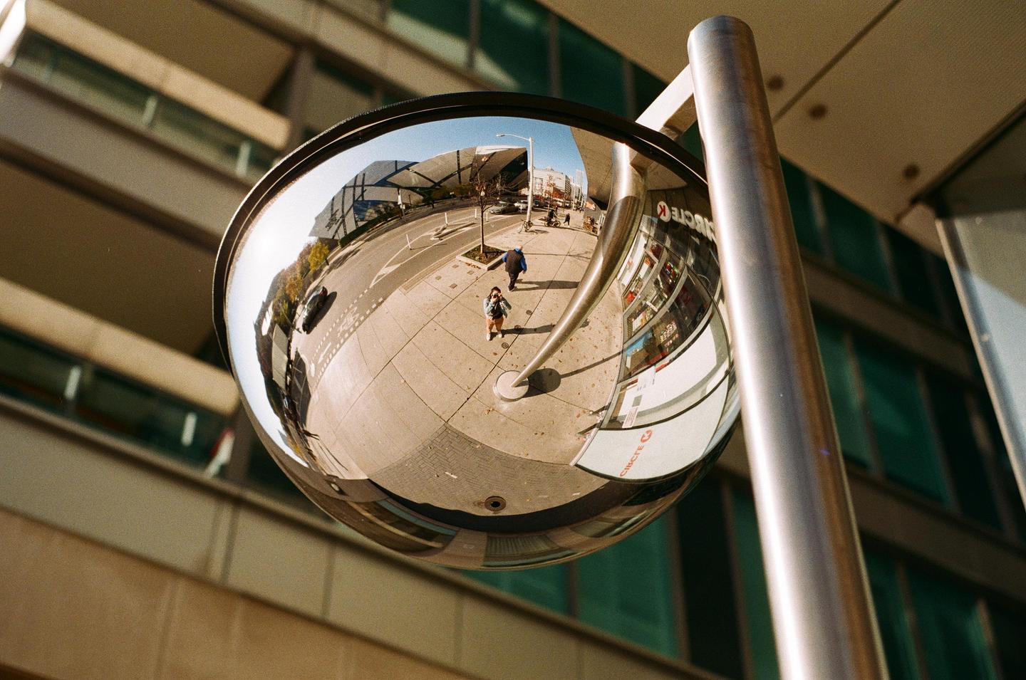 A convex mirror reflects a street scene with a woman and a man walking on a sidewalk.