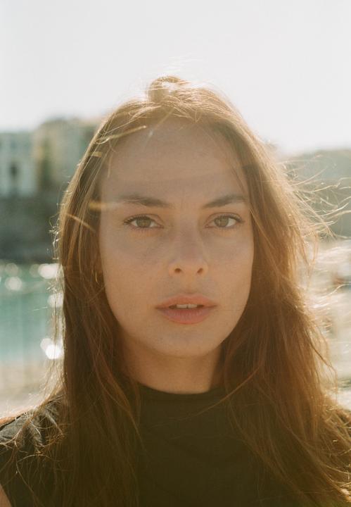 A woman with long hair looks directly at the viewer, with a body of water and buildings in the background.