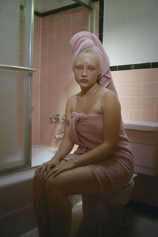 A woman wrapped in a pink towel sits on a toilet in a pink tiled bathroom.
