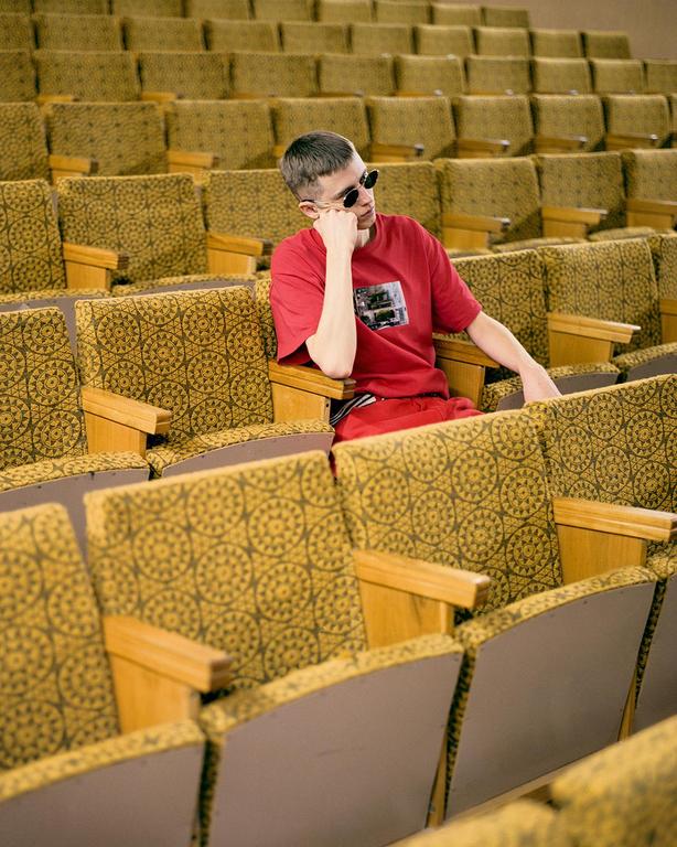A man wearing sunglasses and a red shirt sits alone in an auditorium.