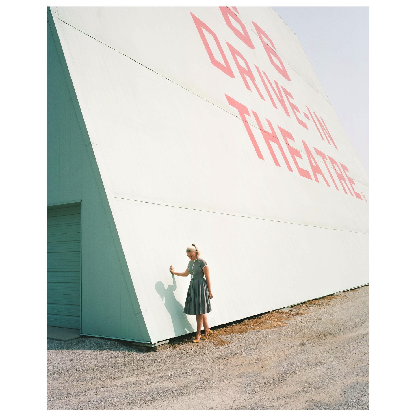 A woman in a gray dress stands next to a large white drive-in theater screen with red lettering.