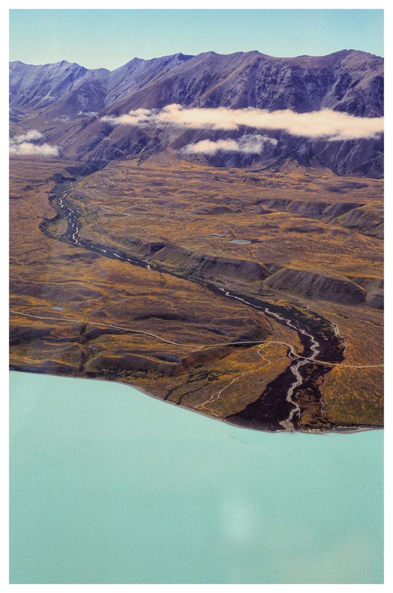 An aerial landscape shows a winding river flowing through a valley with mountains in the background and a large body of water in the foreground.