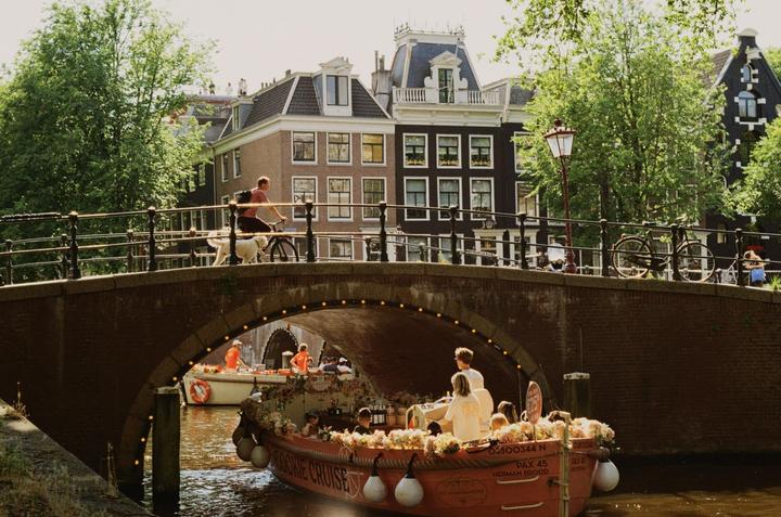 A man rides a bicycle with a dog across a brick bridge over a canal with boats and buildings in the background.